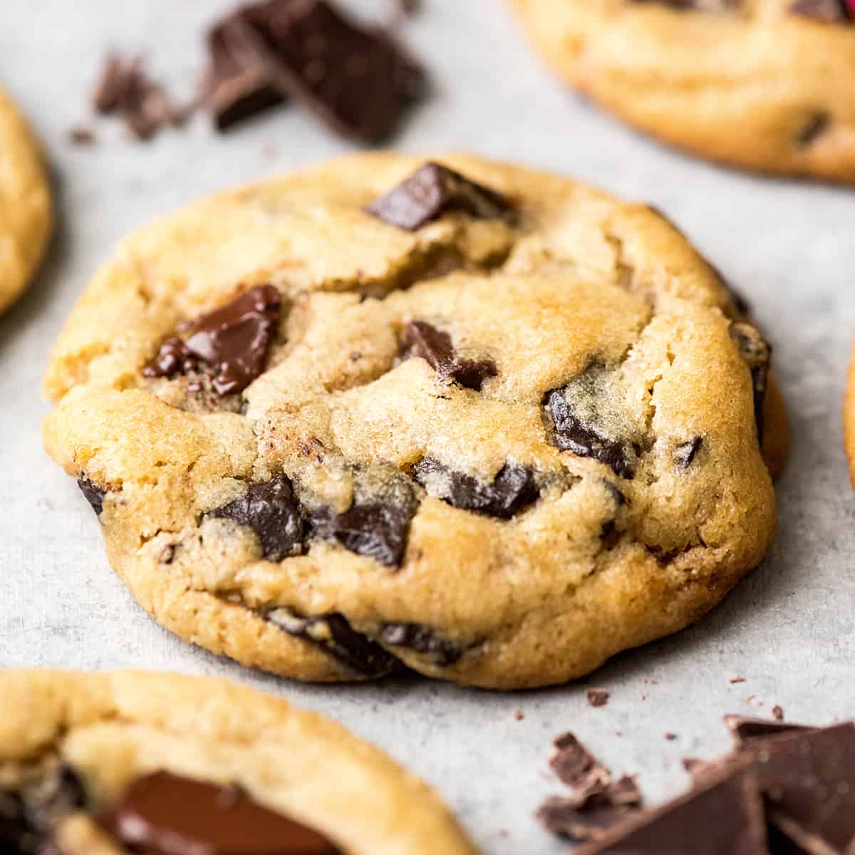 This is the best chocolate chip cookie recipe ever. No funny ingredients, no chilling time, etc. Just a simple, straightforward, amazingly delicious, doughy yet still fully cooked, chocolate chip cookie that turns out perfectly every single time! #cookies #chocolatechipcookies #baking #recipe #chocolate side view of the best chocolate chip cookie recipe ever one cookie with melty chocolate chips surrounded by three other cookies and chopped chocolate