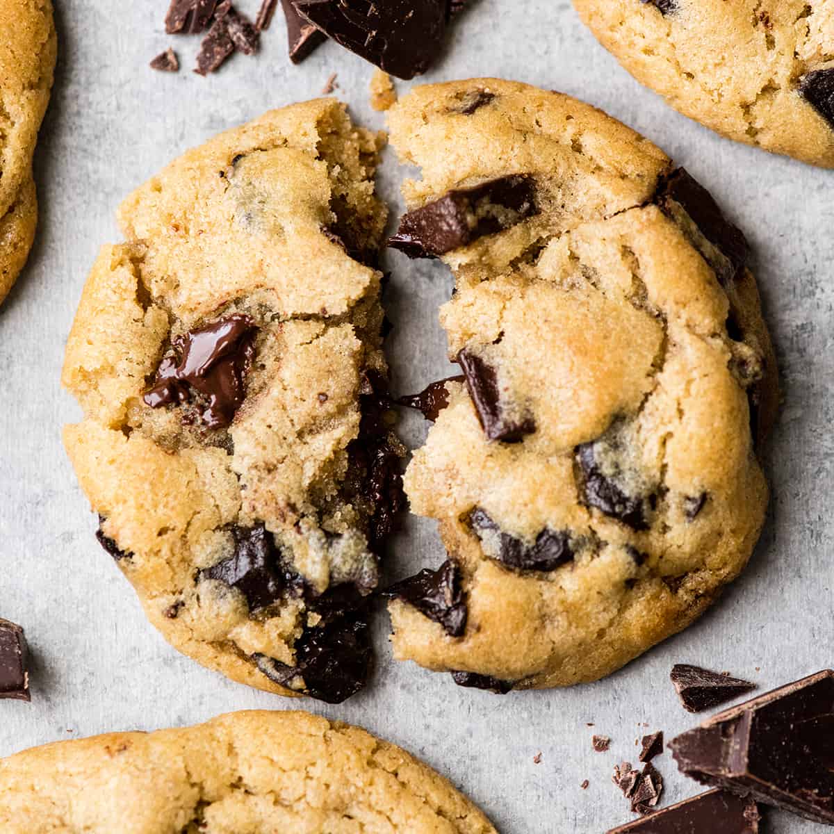 This is the best chocolate chip cookie recipe ever. No funny ingredients, no chilling time, etc. Just a simple, straightforward, amazingly delicious, doughy yet still fully cooked, chocolate chip cookie that turns out perfectly every single time! #cookies #chocolatechipcookies #baking #recipe #chocolate Overhead view of one chocolate chip cookie torn in half with melty chocolate chips surrounded by three other cookies and chopped chocolate