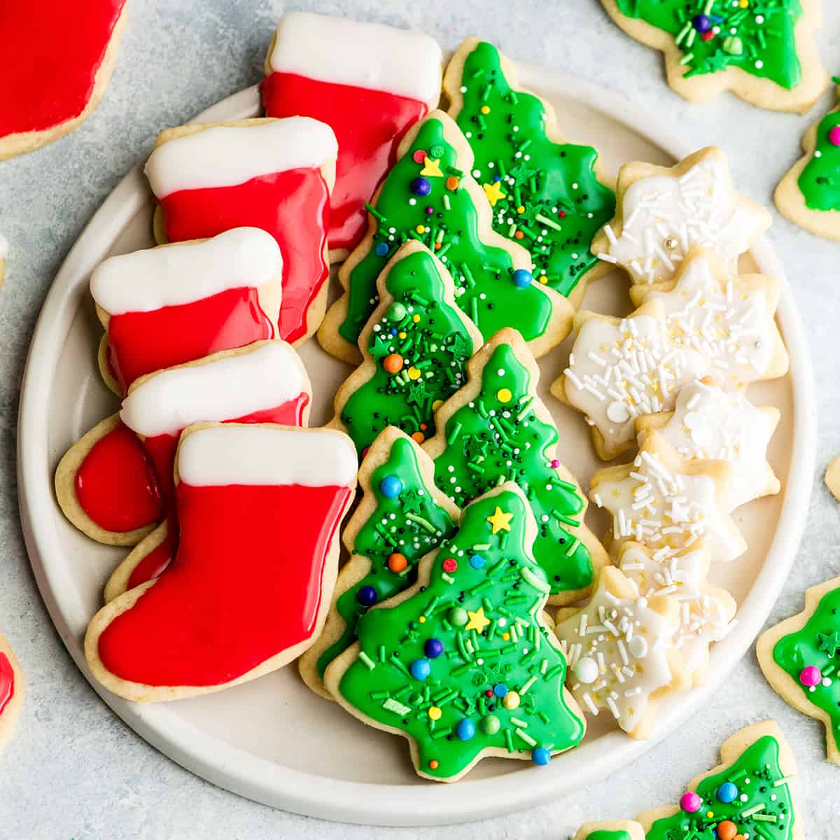 overhead view of a white, round plate with three rows of sugar cookies on it. The left row is five red and white stockings, the middle row is six green Christmas trees with sprinkles and the left row is seven small white stars with sprinkles. 