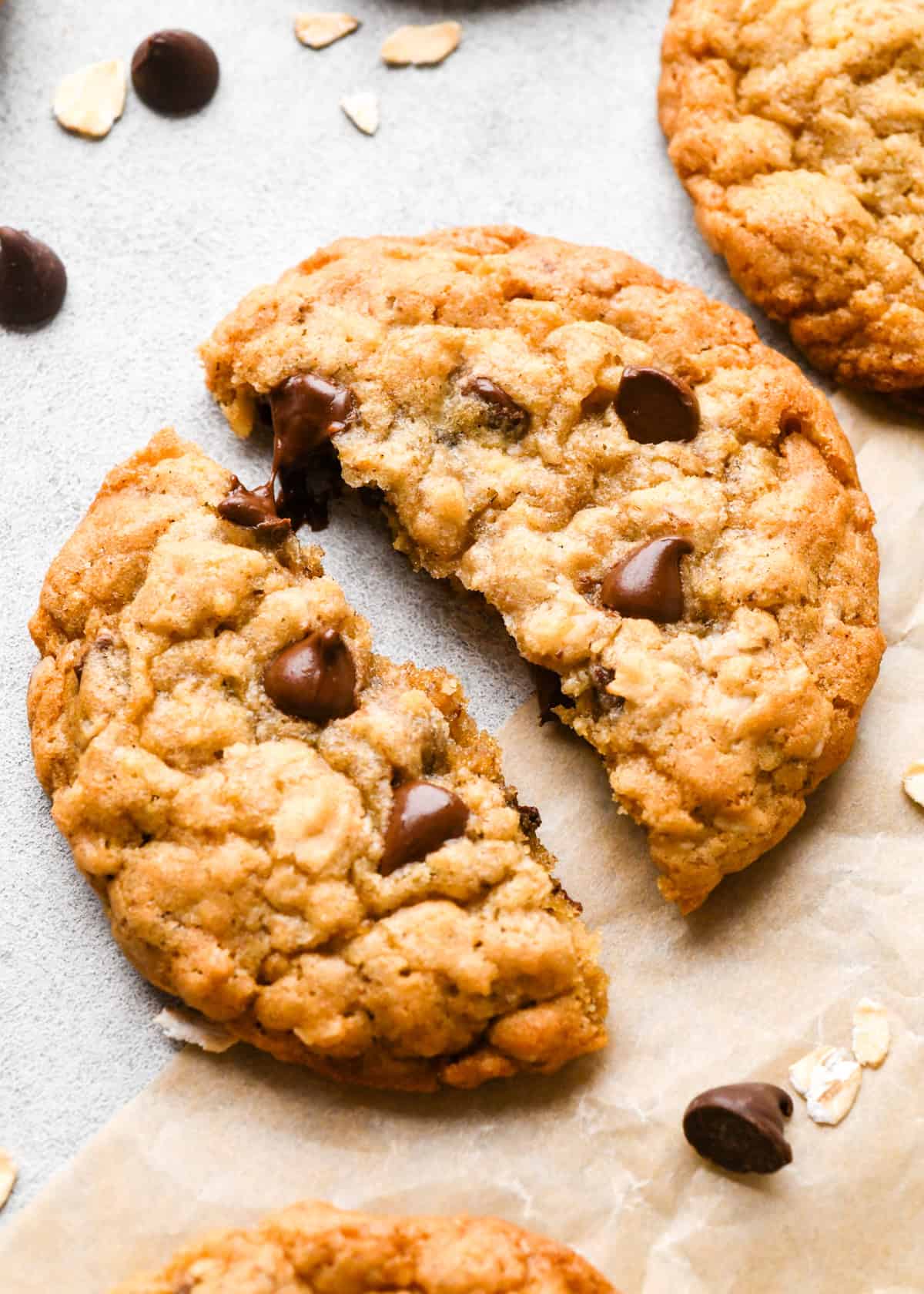 3 Oatmeal Chocolate Chip Cookies on parchment paper, one cut in half, surrounded by raw oats and chocolate chips. 