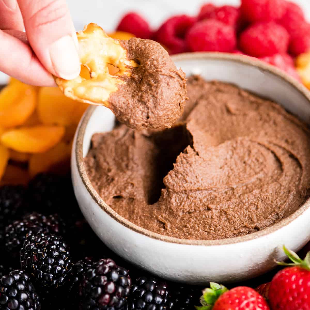Front view of a hand dipping a pretzel chip into a bowl of Chocolate Hummus