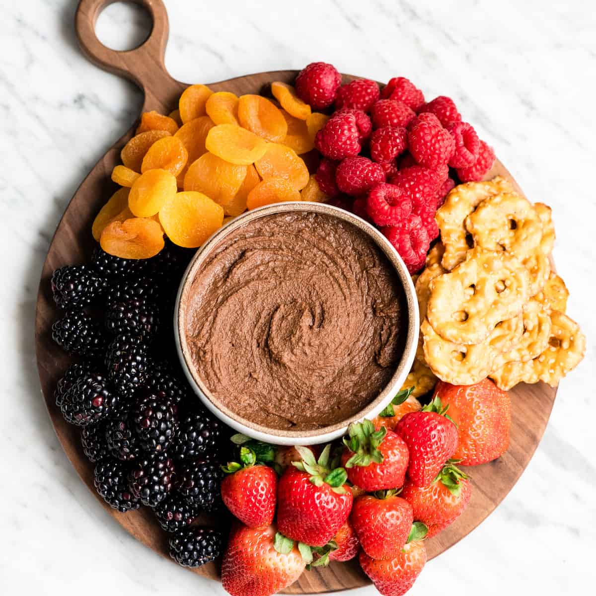 Healthy Chocolate Hummus Recipe is easy to make and ready in 5 minutes! It's a nutritious dip, appetizer, dessert, snack or spread that everyone in my family, including my kids, loves. This dessert hummus recipe is gluten-free, dairy-free, has no refined sugar & it's vegan! #hummus #chocolatehummus #vitamix #desserthummus #vegan Overhead view of Chocolate Hummus in a bowl surrounded by fruits and pretzels to dip in it