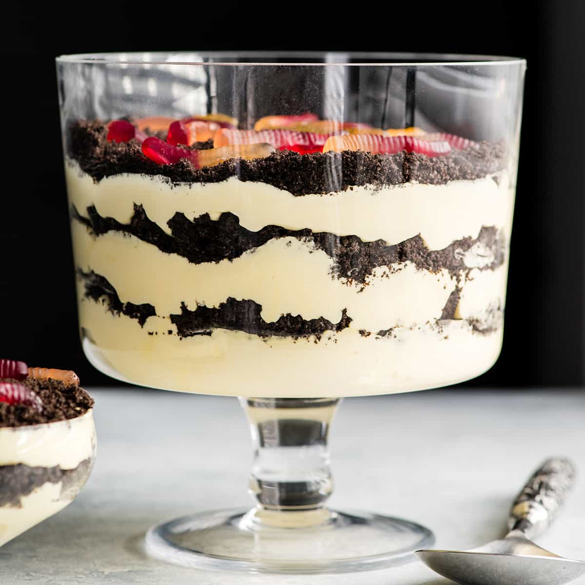 This Easy Dirt Cake Recipe (Oreo Dirt Pudding) is one of our favorite no-bake desserts! Crushed Oreos are layered between a french vanilla pudding mixture for a kid-friendly, make-ahead dessert recipe perfect for any potluck or BBQ! Add gummy worms for a whimsical touch! #dirtcake #dirtpudding #oreos #oreopudding #nobake #dessert #potluck #bbq #summer Front view of this Oreo Dirt Cake Recipe in a glass trifle dish with three layers of pudding and three layers of crushed Oreos with gummy worms on top and a spoon and a single-serve dish of Oreo Dirt Cake next to it