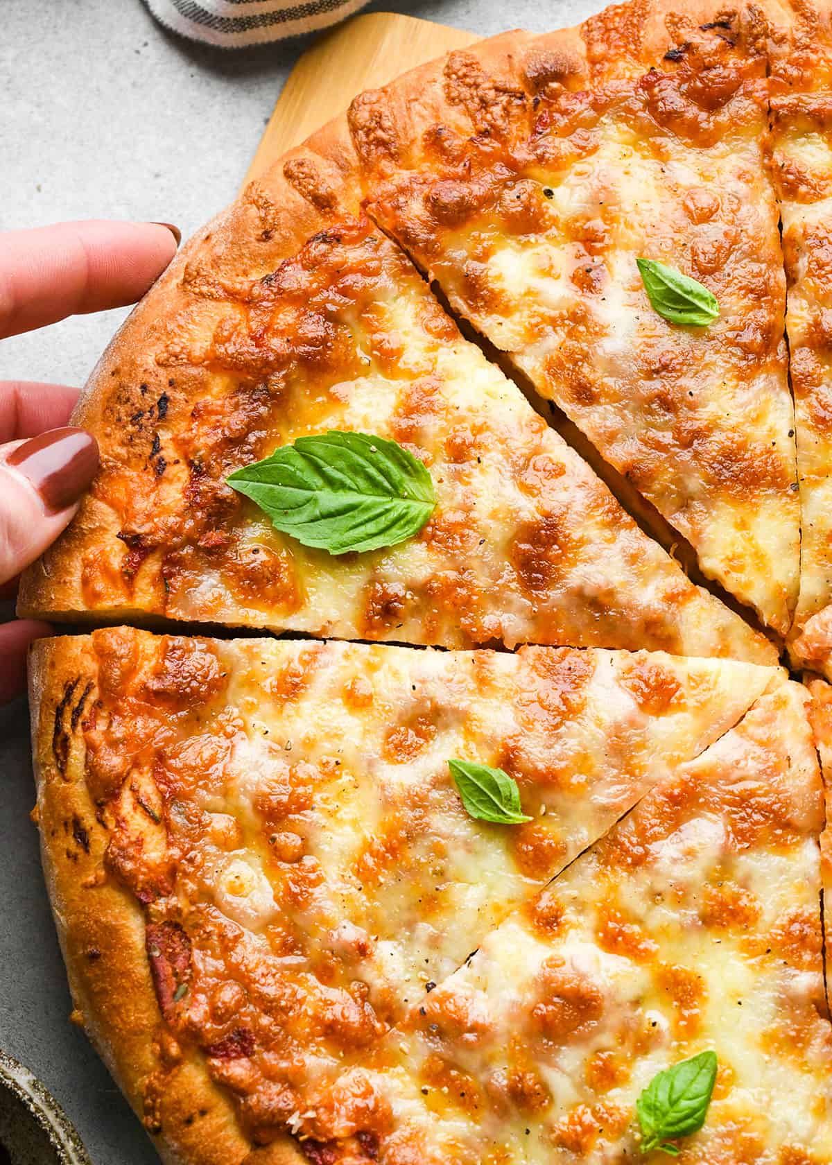 baked pizza made with homemade pizza dough cut into triangles, garnished with basil. A hand is taking a piece of pizza from the pizza