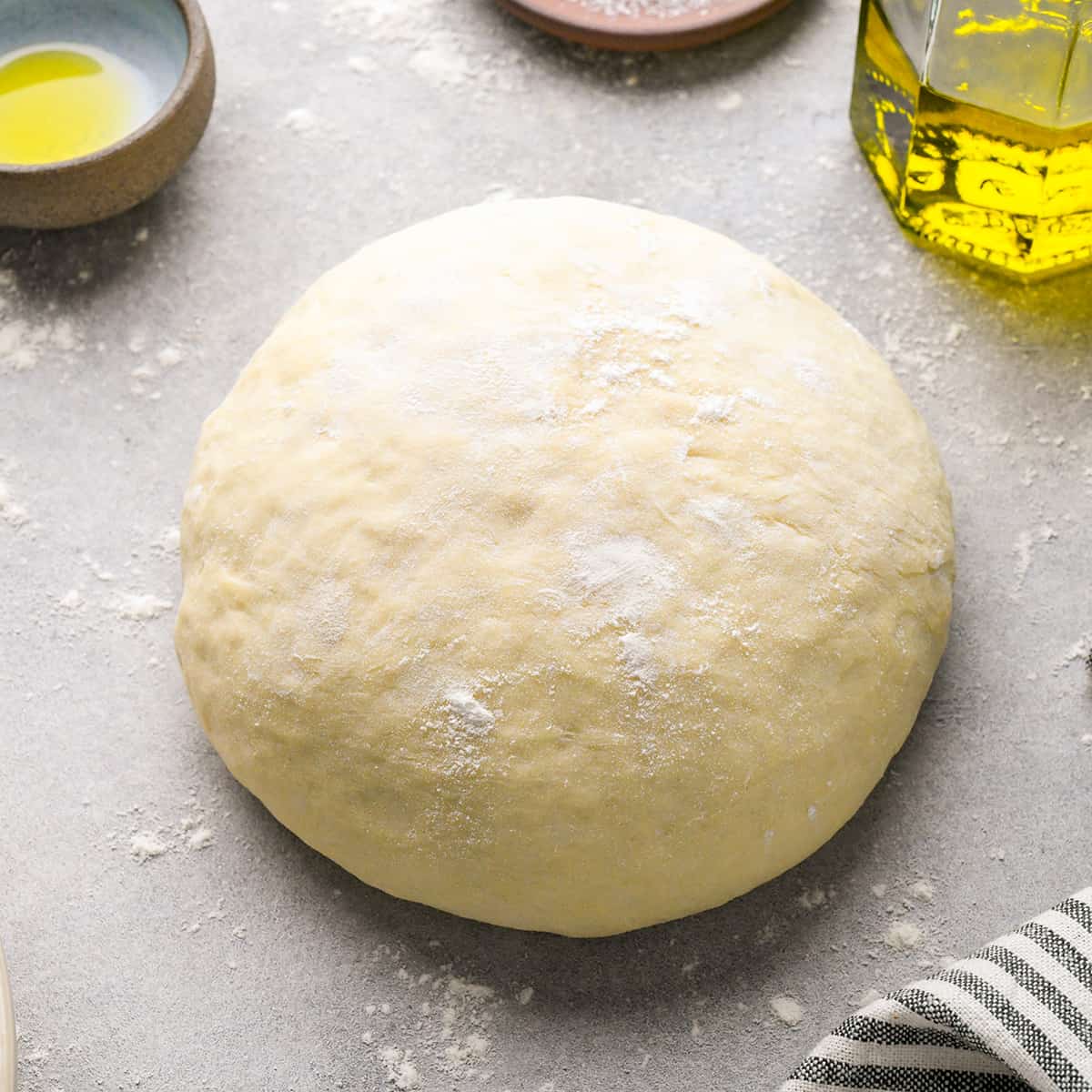 a ball of homemade pizza dough dusted with flour on a gray floured surface with olive oil and a striped towel nearby