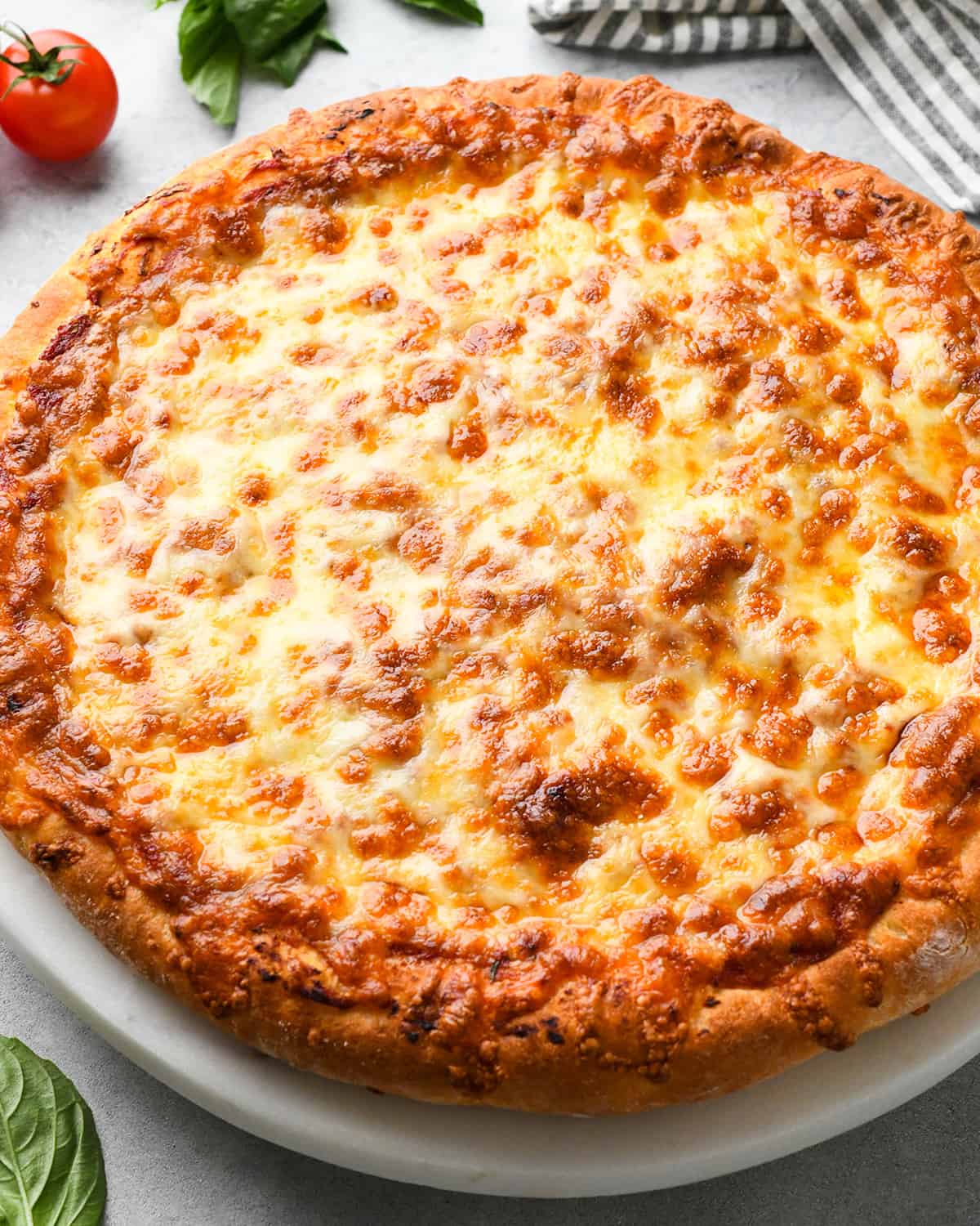 a baked pizza with homemade pizza dough on a serving plate with tomato, basil and a striped towel nearby