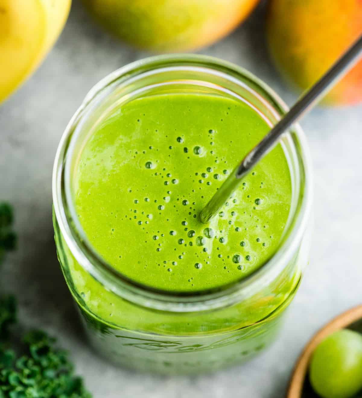 overhead view of a green smoothie in a large glass mason jar with a metal straw, surrounded with kale, bananas, mangoes and green grapes
