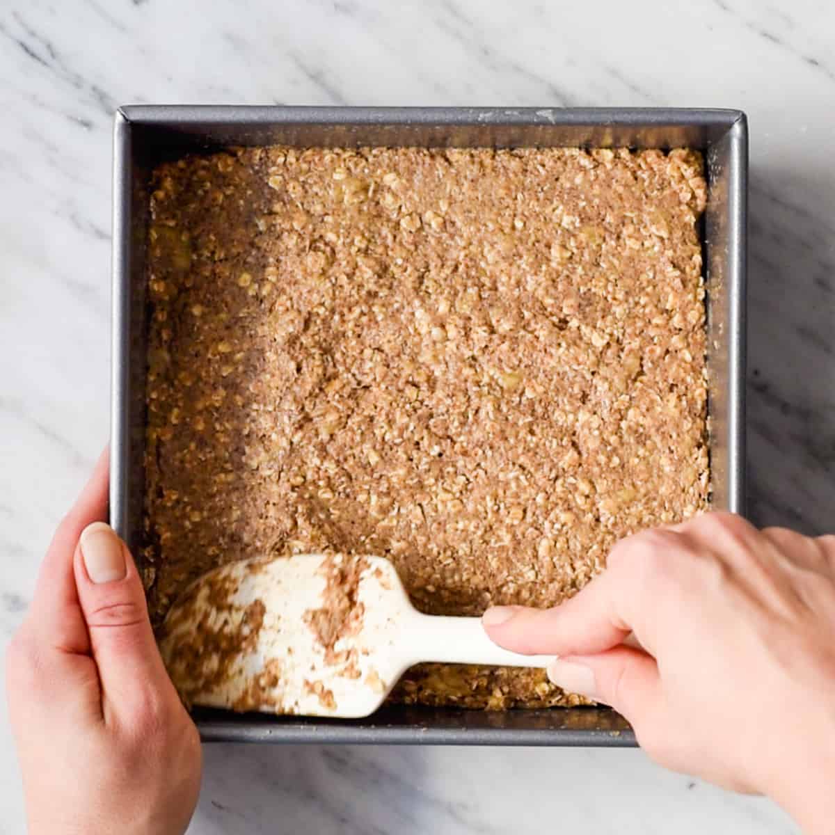 overhead view of hands spreading the breakfast bar batter into a square pan with a white spatula before baking. 