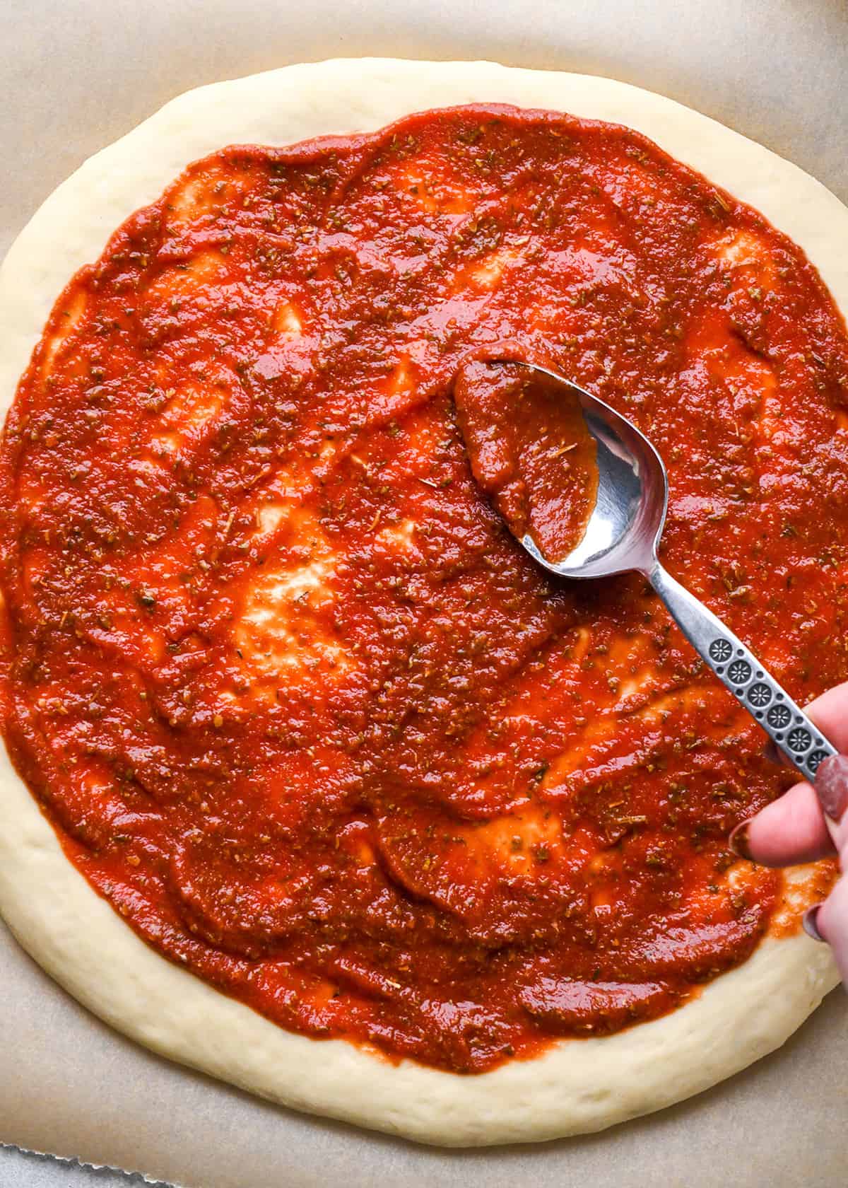 pizza sauce being spread on homemade pizza dough with a silver metal spoon. the dough is on brown parchment paper