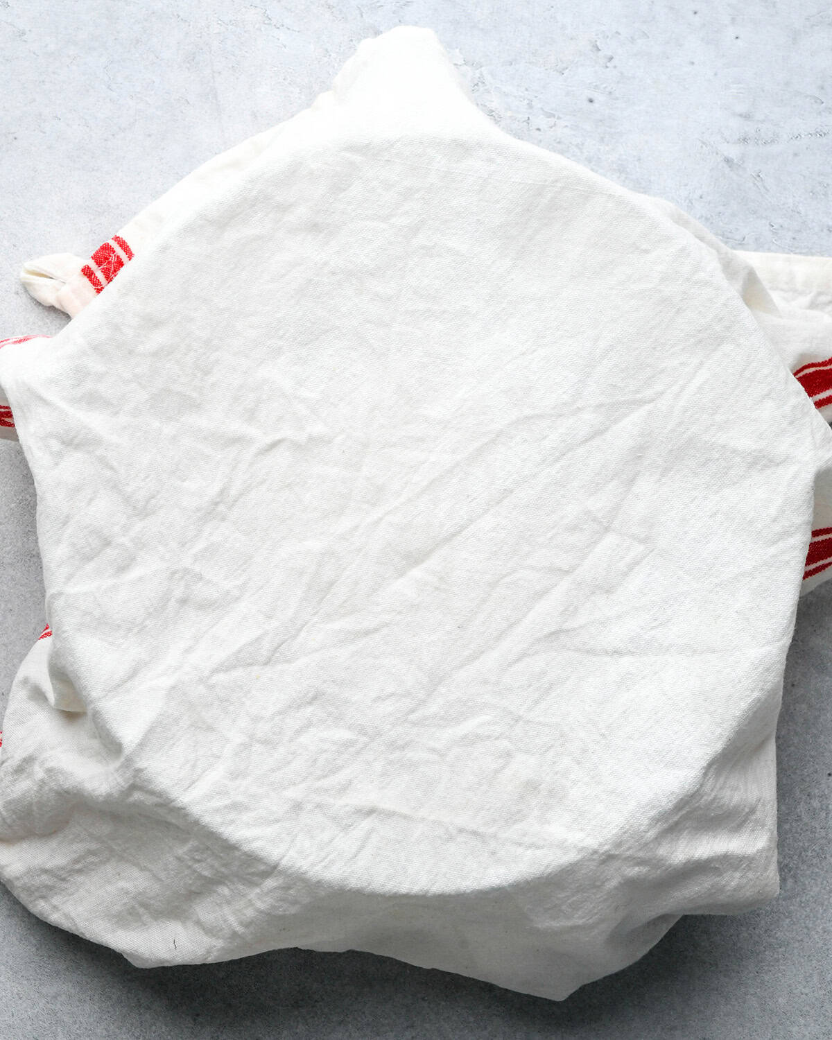 pizza dough in a bowl on a gray surface covered by a damp white and red tea towel 