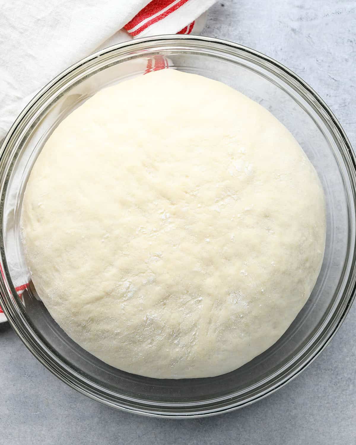 risen pizza dough in a glass bowl on a gray surface with a white and red tea towel in the top left corner 
