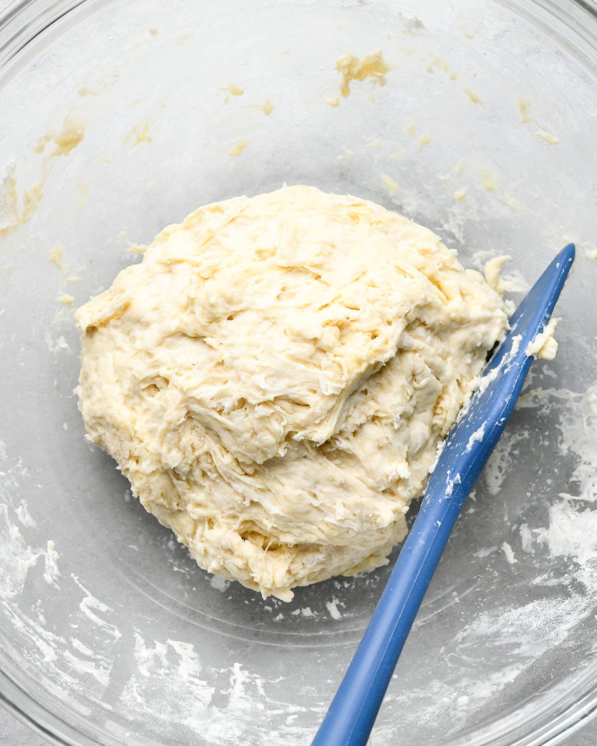a ball of pizza dough in a glass bowl with a blue spatula on a gray surface