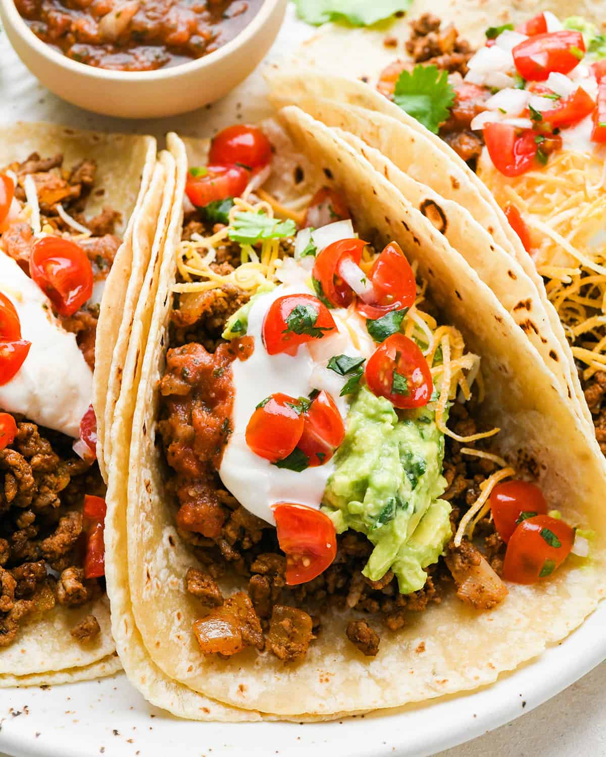 3 Ground Beef Tacos on a plate with sour cream, cheese, guacamole and tomatoes