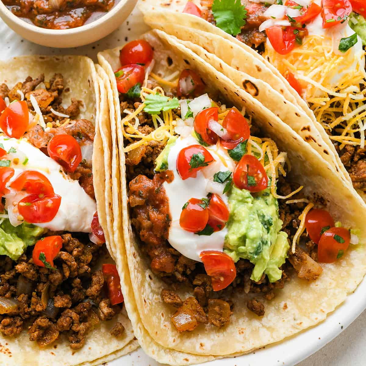 3 Ground Beef Tacos on a plate with sour cream, cheese, guacamole and tomatoes