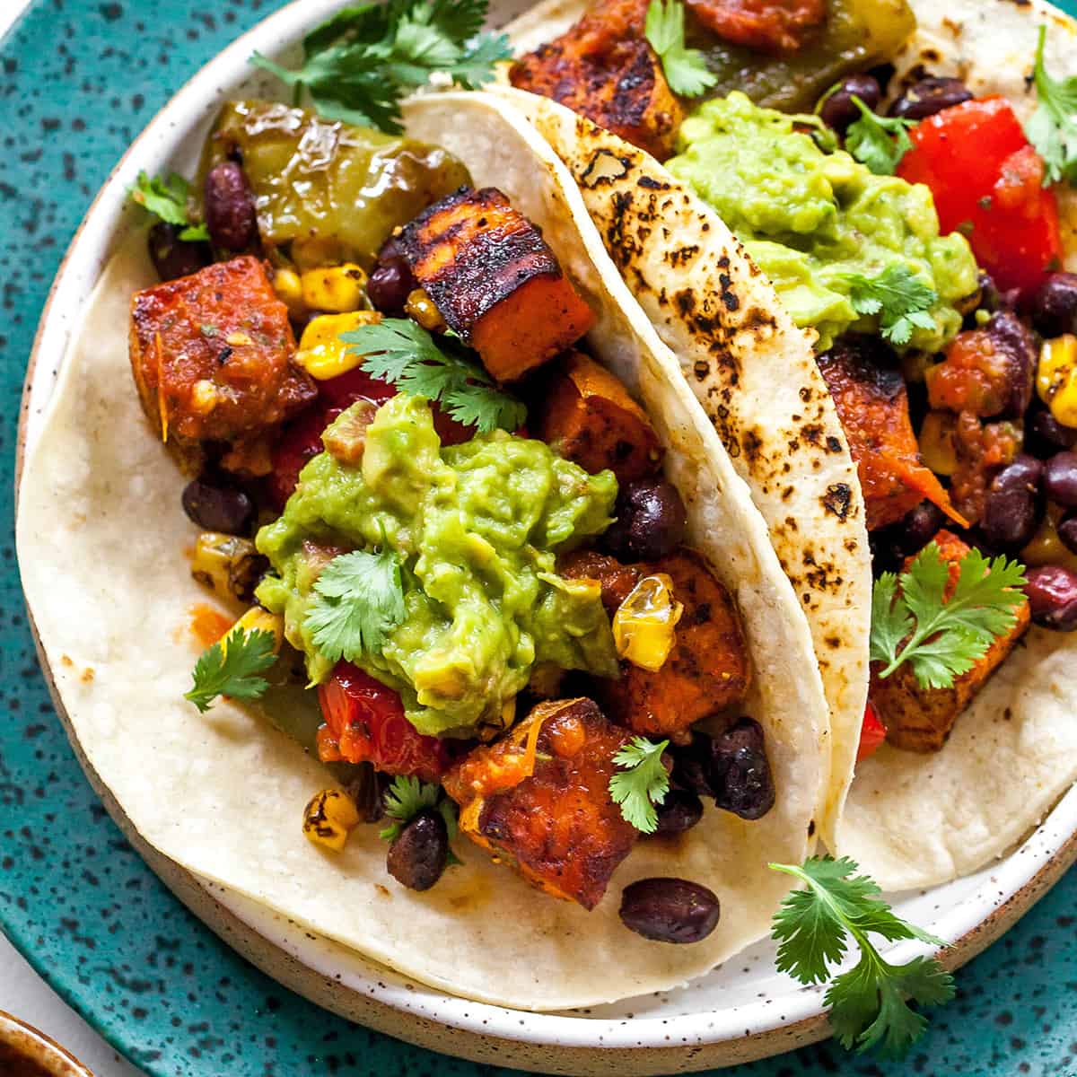 These healthy black bean sweet potato tacos are the best vegetarian tacos ever. Sweet potatoes and vegetables are perfectly spiced and roasted, then tossed with a honey, lime cilantro mixture for the most flavorful and delicious tacos. Taco night isn't complete without this sweet potato taco recipe! two black bean sweet potato tacos on a plate with guacamole and cilantro