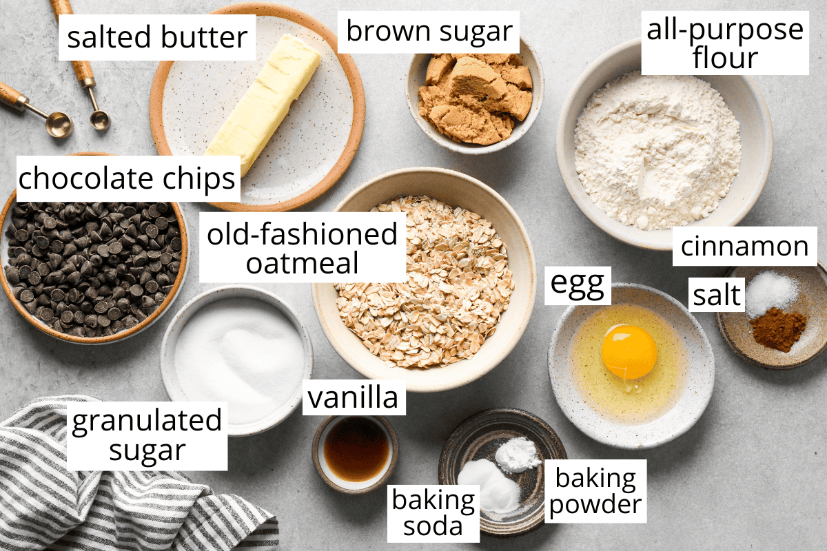 overhead photo of the labeled ingredients in this Oatmeal Chocolate Chip Cookies recipe like butter, oatmeal, flour, brown and white sugar, chocolate chips, egg, cinnamon, etc. 